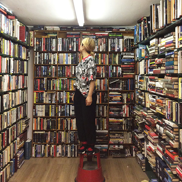 イギリス・ロンドンの古本屋さんで、3面の本棚に囲まれて踏み台の上に立つアーティスト・norahiさん